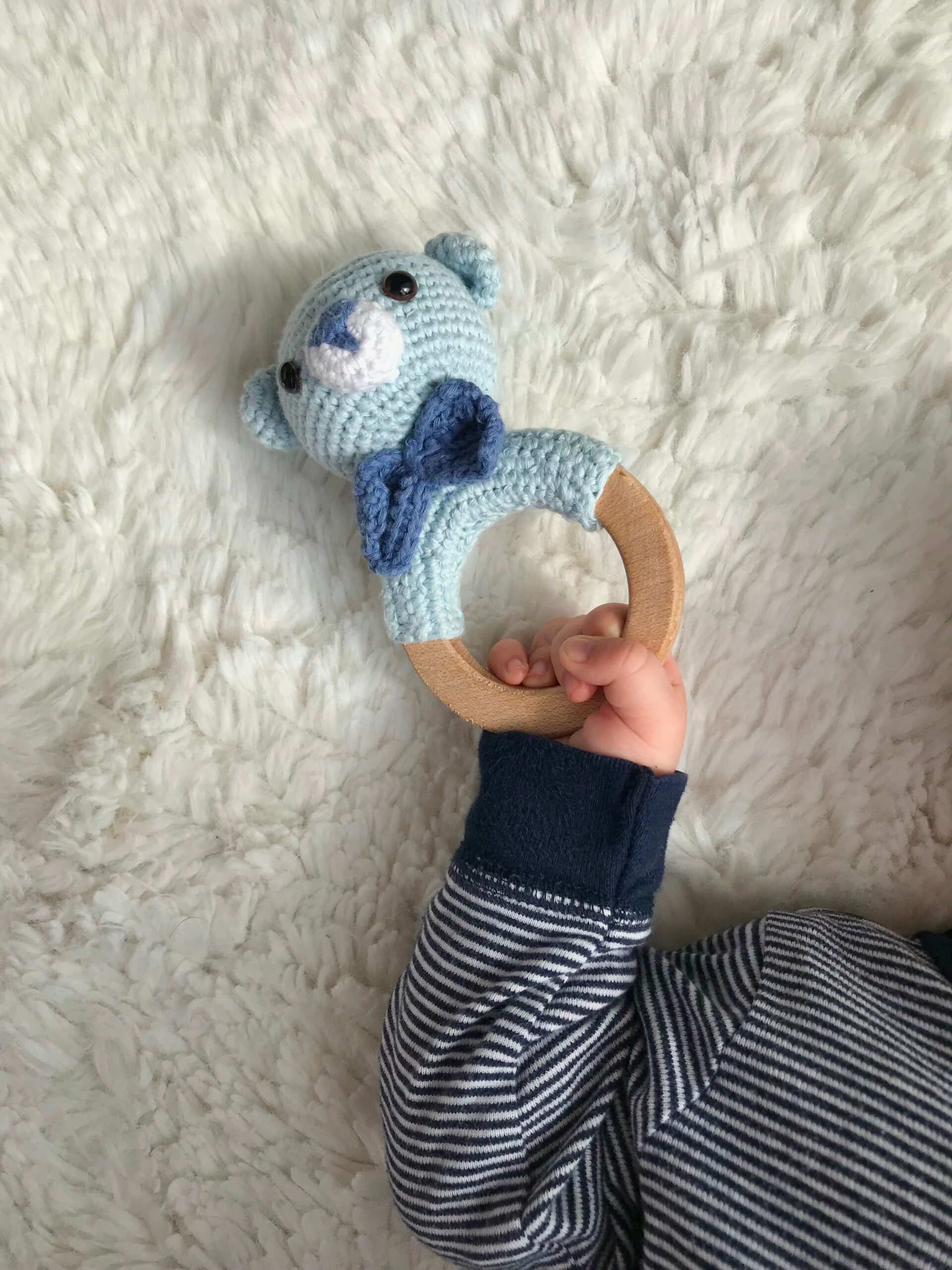 A cute baby holds a knitted blue teddy bear ring on a soft fur carpet. Perfect for childcare themes.